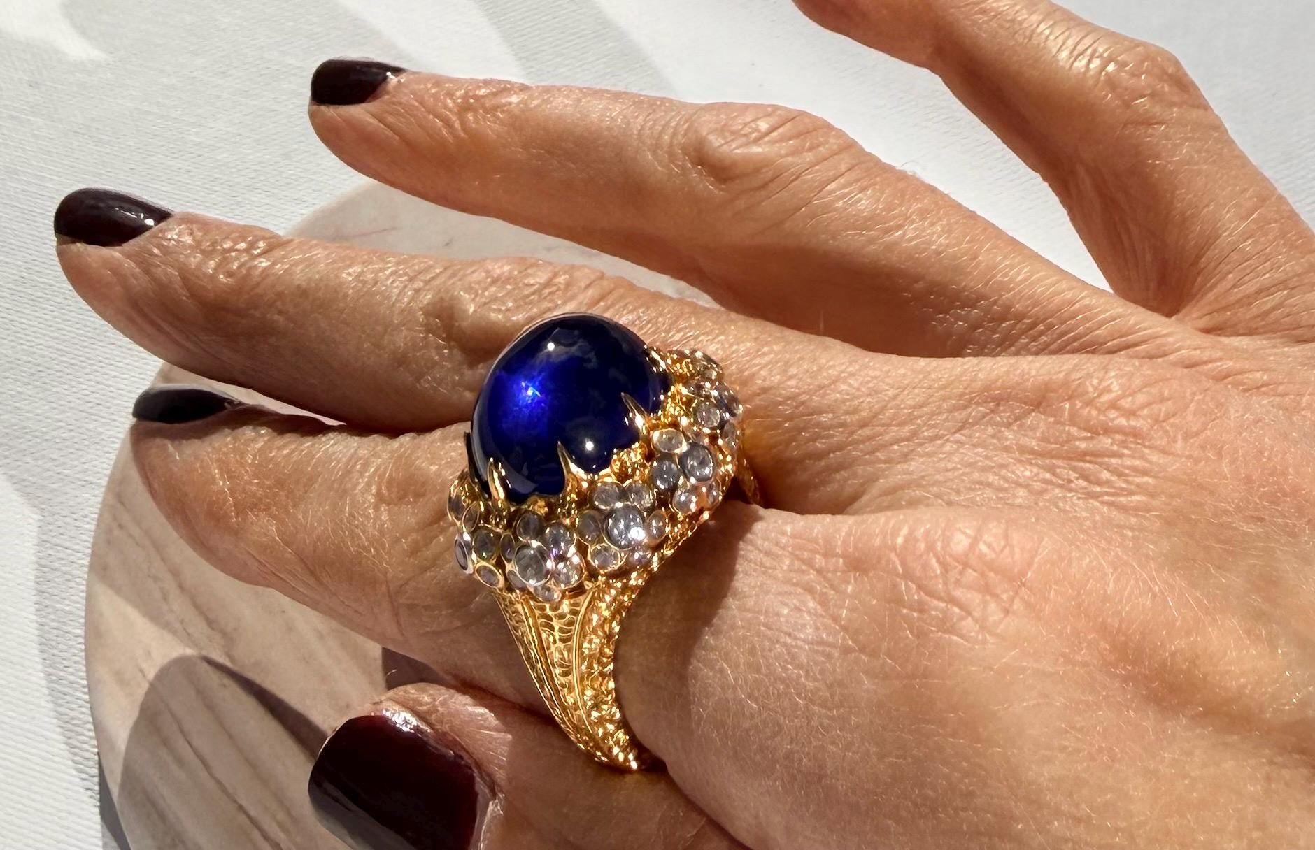 Fine jewelry yellow gold ring featuring a large deep blue cabochon sapphire and a sparkling diamond halo worn on a hand with dark burgundy nail polish over an intricately textured gold shank