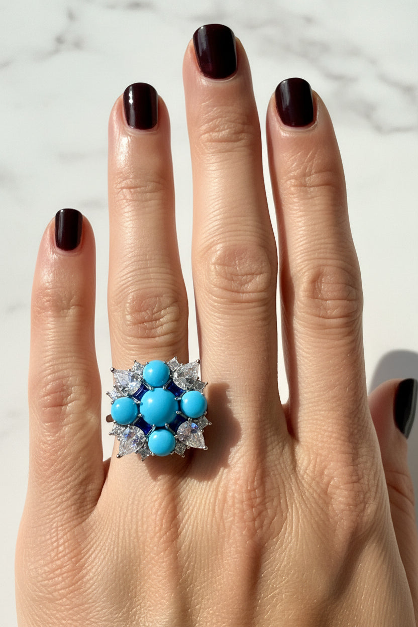 Jewelry photography of a statement cocktail ring on a hand with dark burgundy nails featuring vivid sky-blue turquoise cabochons, sparkling pear-cut stones, and deep navy accents set against a white marble backdrop