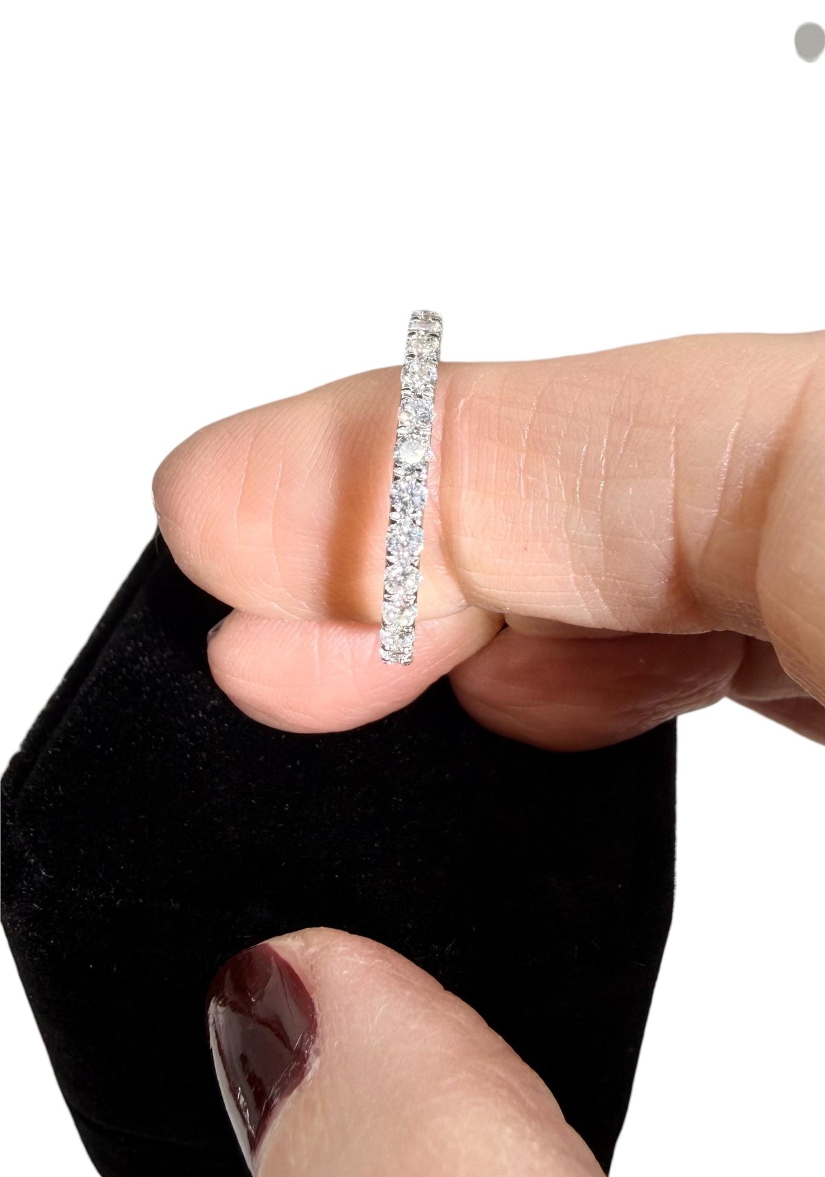 Photograph of a diamond eternity band held between fingers above a black velvet box featuring brilliant round-cut diamonds, a slim white gold setting, and a thumb with glossy maroon nail polish
