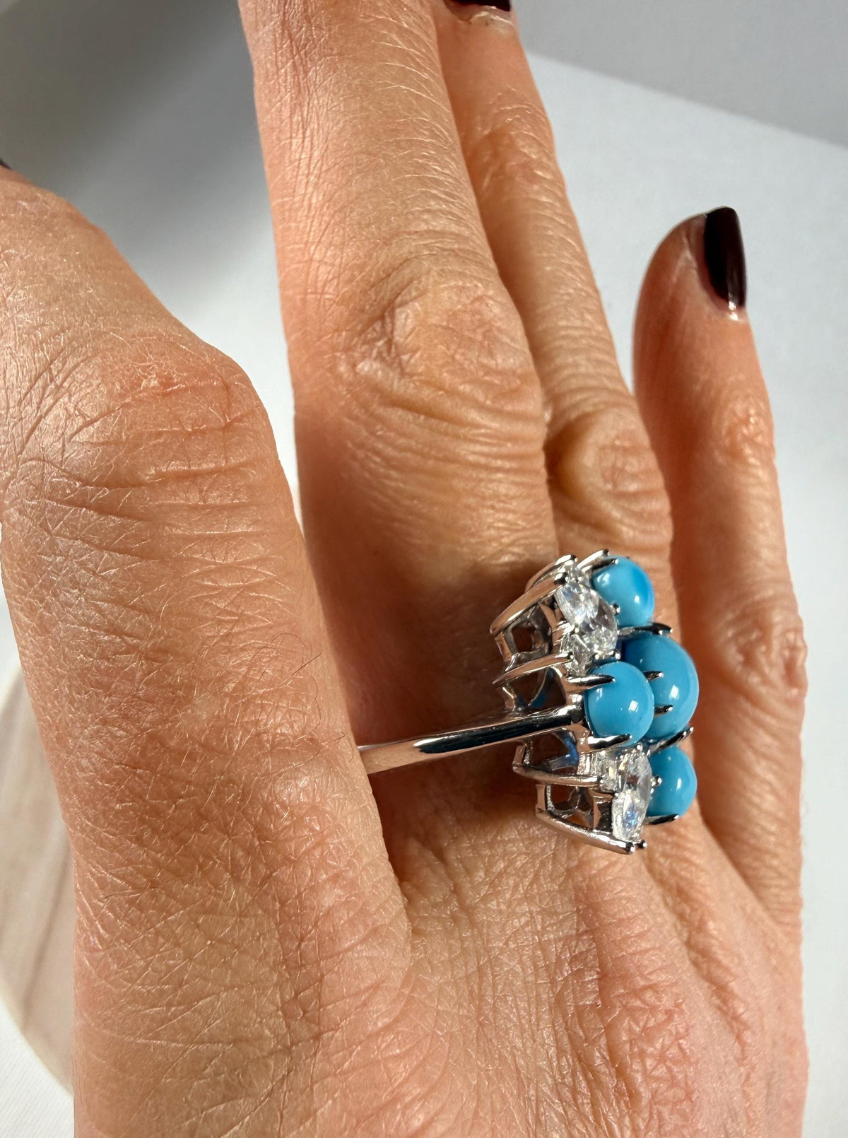 Photograph of a silver-toned cocktail ring featuring five vibrant blue turquoise cabochons and sparkling clear accent stones worn on a ring finger with dark plum nail polish