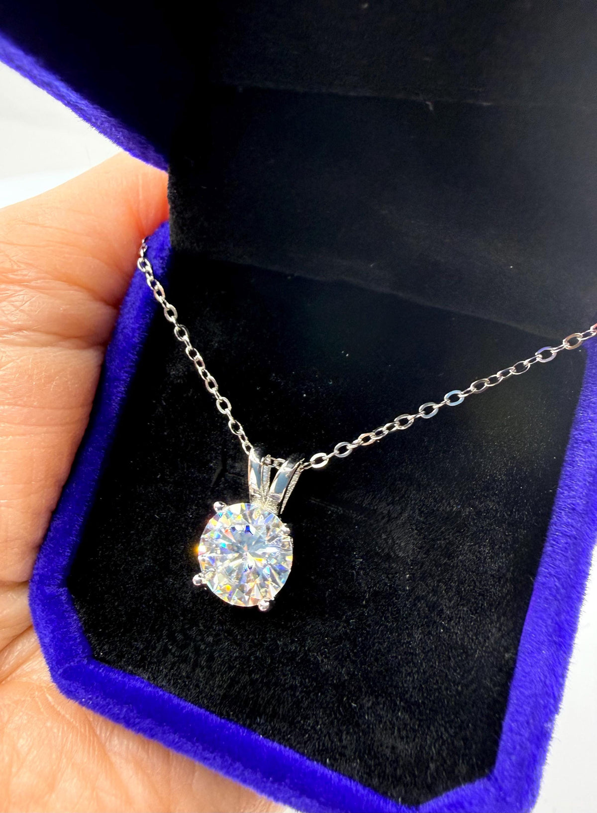 Photograph of a brilliant round-cut solitaire pendant necklace resting in a royal blue velvet box held by a hand, showcasing a sparkling multifaceted stone with prismatic light reflections and a delicate silver chain