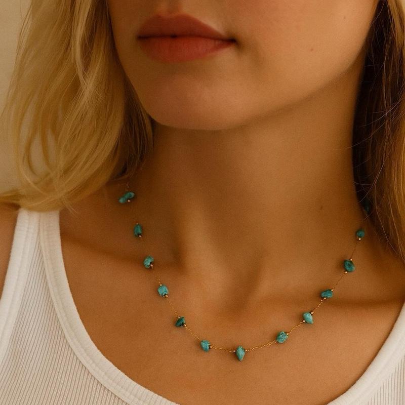 Photograph of a blonde woman's neck showcasing a delicate gold chain necklace with organic turquoise beads worn over a white ribbed tank top with a hint of red lipstick