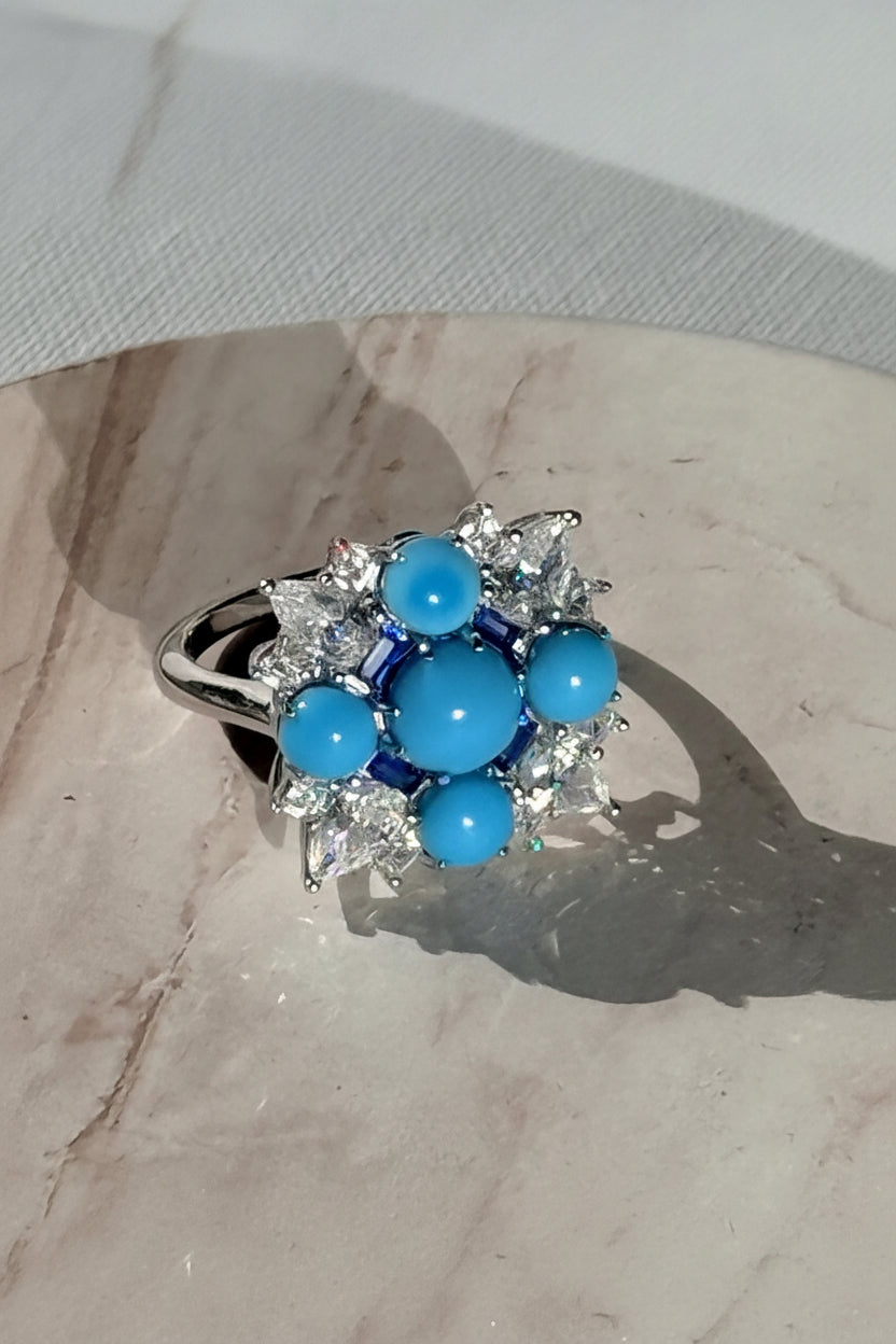Fine jewelry ring resting on a beige marble surface featuring five vibrant turquoise cabochons in a cross formation surrounded by brilliant pear-cut clear stones and small blue baguette accents