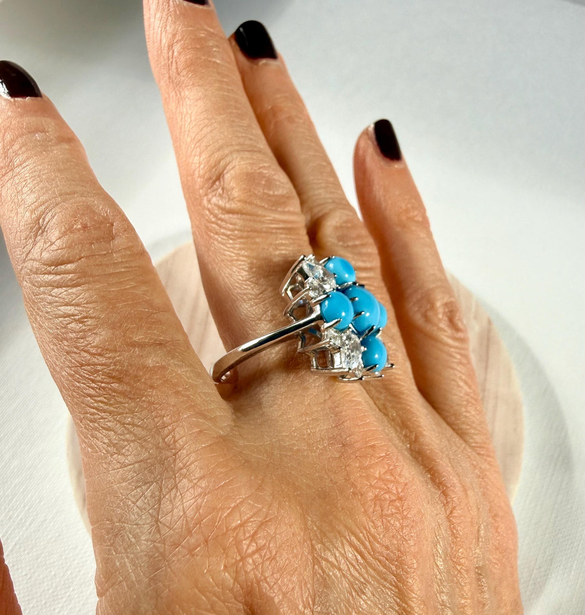 Photograph of a sterling silver cocktail ring featuring vibrant blue turquoise cabochons and sparkling clear gemstones worn on a hand with dark polished nails