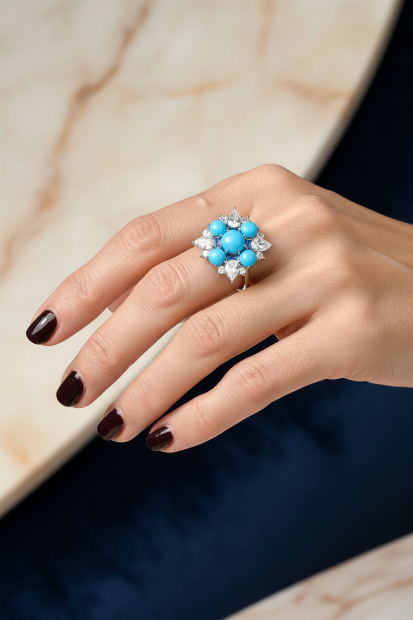 Photograph of a hand wearing a large statement ring featuring vibrant turquoise cabochons and shimmering pear-shaped diamonds, detailed with dark burgundy-black nail polish and set against a contrasting marble and navy blue background