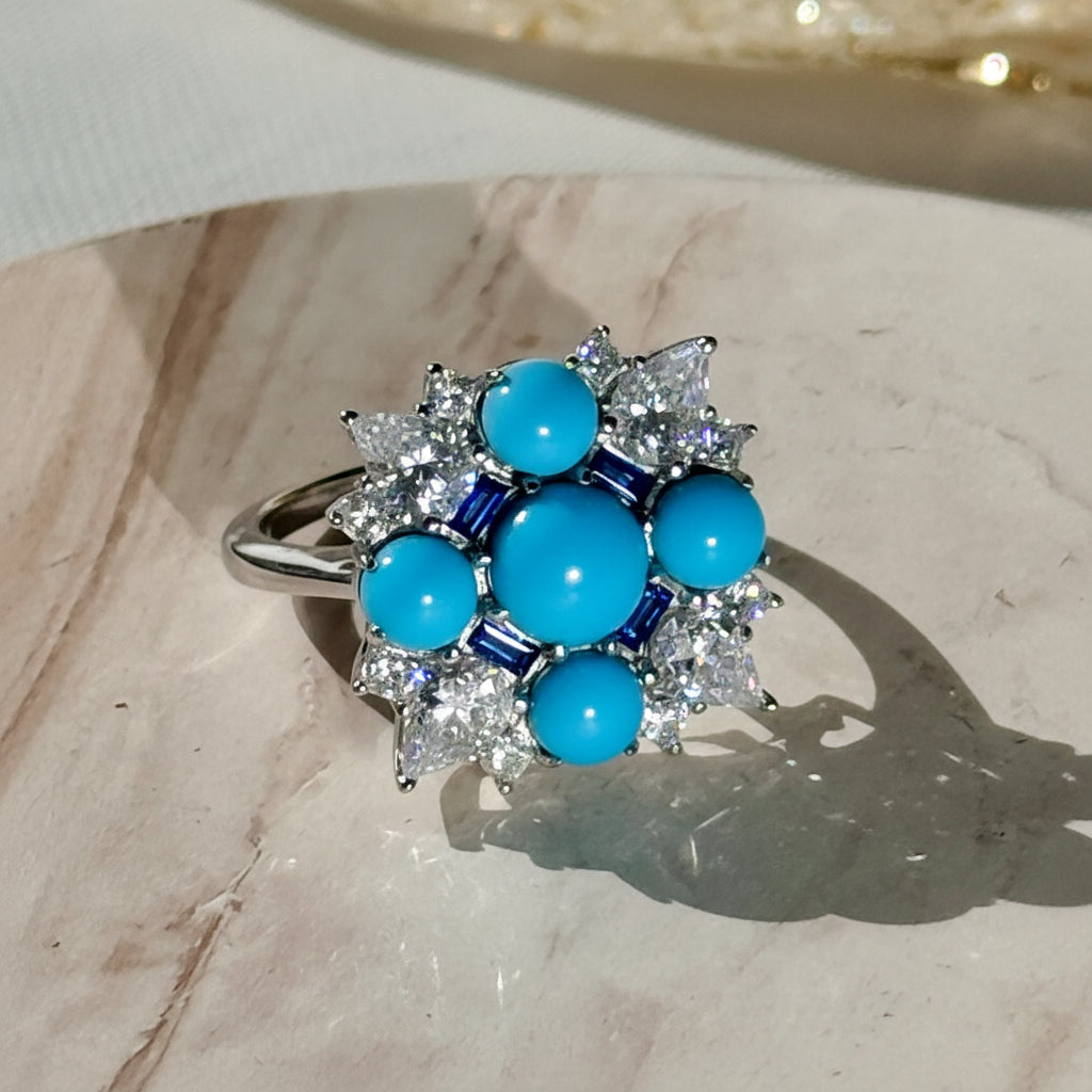 Fine jewelry silver-toned ring featuring five vivid blue turquoise cabochons surrounded by sparkling white pear-cut diamonds and dark blue baguette sapphire accents resting on a light-colored marbled surface