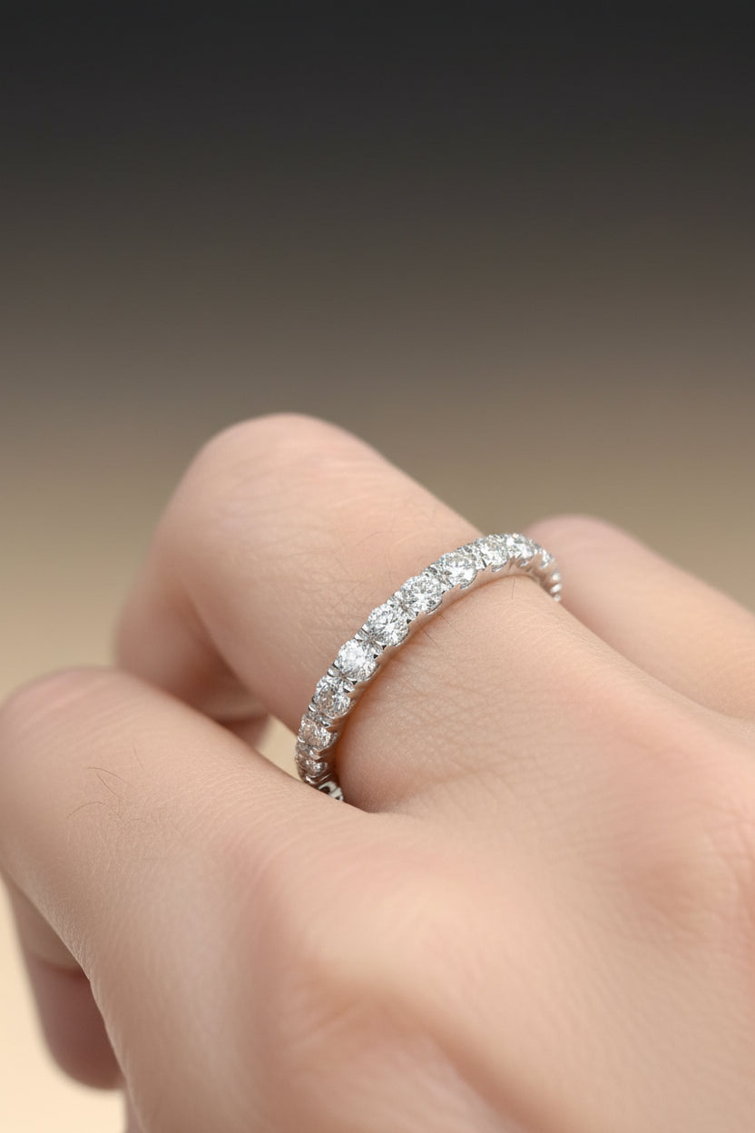 Macro photograph of a diamond eternity band on a human finger highlighting sparkling round-cut gems and a delicate white gold setting against a soft tan backdrop