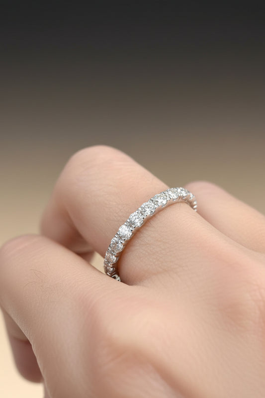 Macro photograph of a diamond eternity band on a human finger highlighting sparkling round-cut gems and a delicate white gold setting against a soft tan backdrop