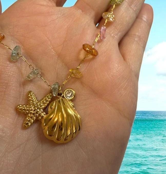 Gold jewelry necklace held in a hand featuring a textured starfish and polished seashell pendant against a tropical turquoise ocean background with multi-colored gemstone beads and a small clear diamond accent