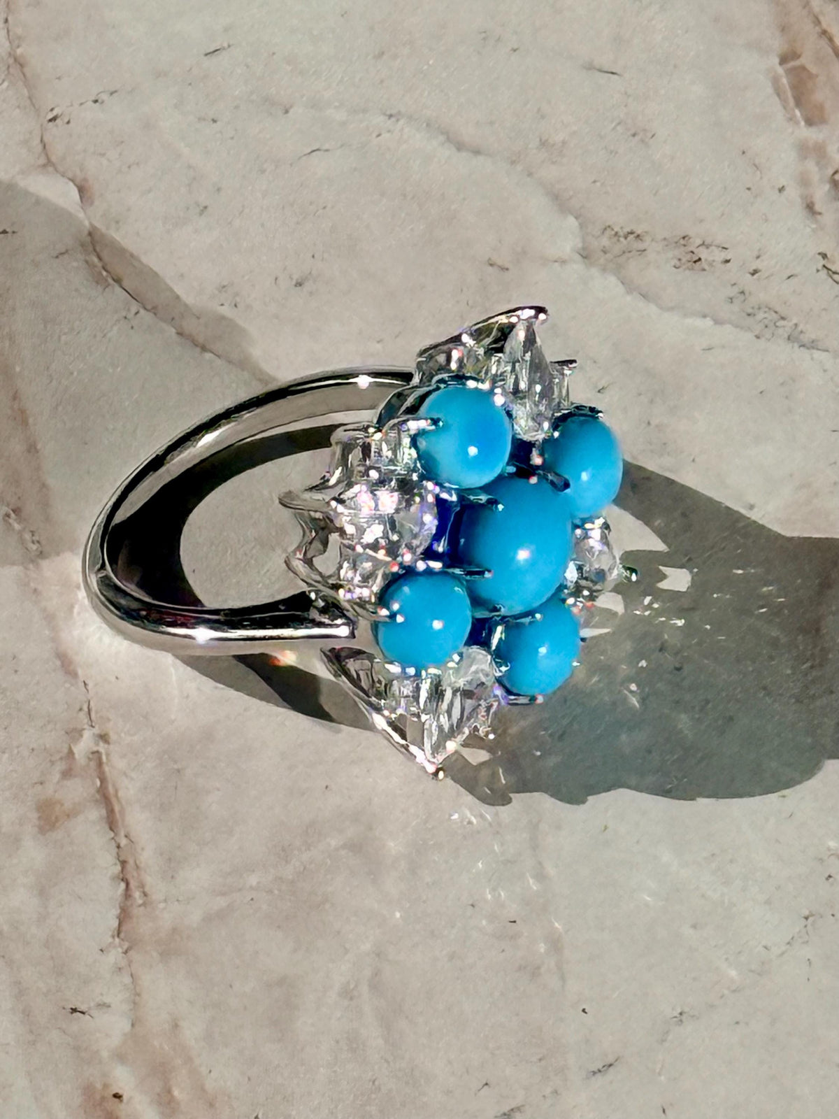 Fine jewelry turquoise and diamond cocktail ring resting on a textured marble surface featuring five vibrant sky-blue cabochons, sparkling marquise-cut diamonds, and a polished white metal band casting a distinct shadow