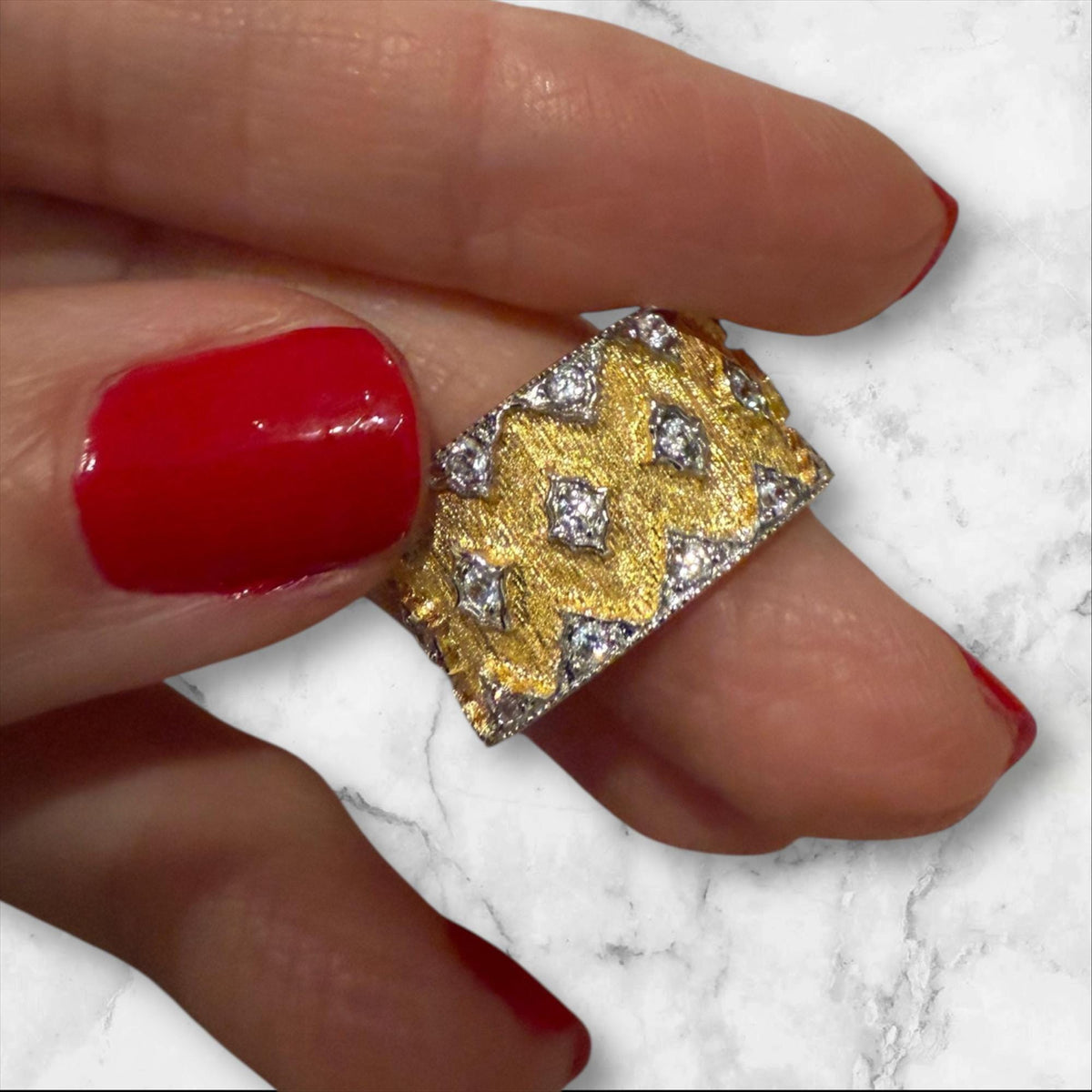 Photograph of a Buccellati two-tone gold and diamond ring held by fingers with vibrant red nail polish against a white marble background featuring hand-engraved rigato texture and star-shaped settings