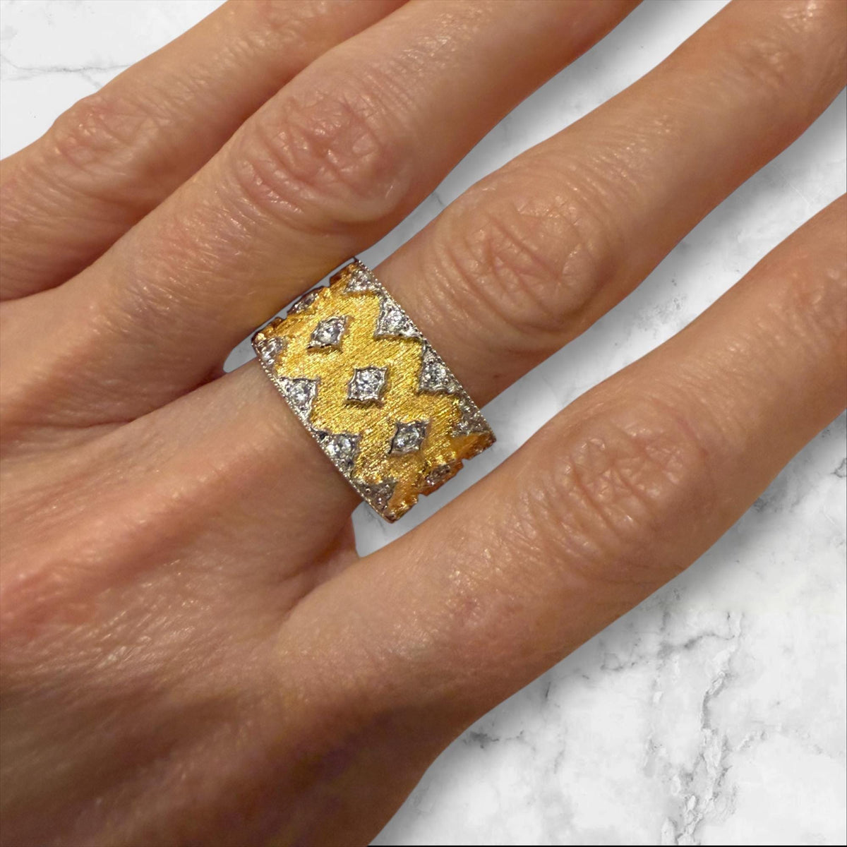 Gold and diamond ring in the Buccellati style worn on a finger over a marble surface featuring yellow gold with rigato engraving, scalloped white gold borders, and star-shaped diamond accents