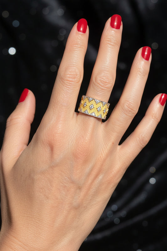 Professional photograph of a hand wearing a wide yellow gold and diamond honeycomb band ring featuring vibrant red manicured nails against a dark shimmering background with soft bokeh light effects