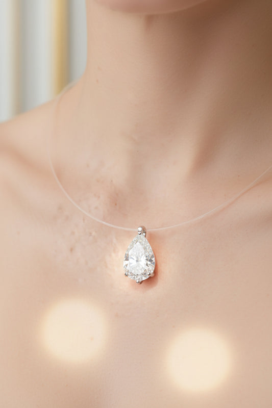 Photograph of a pear-shaped diamond pendant worn on a woman's neck featuring a brilliant teardrop-cut stone suspended on a transparent cord with soft golden bokeh highlights in the foreground