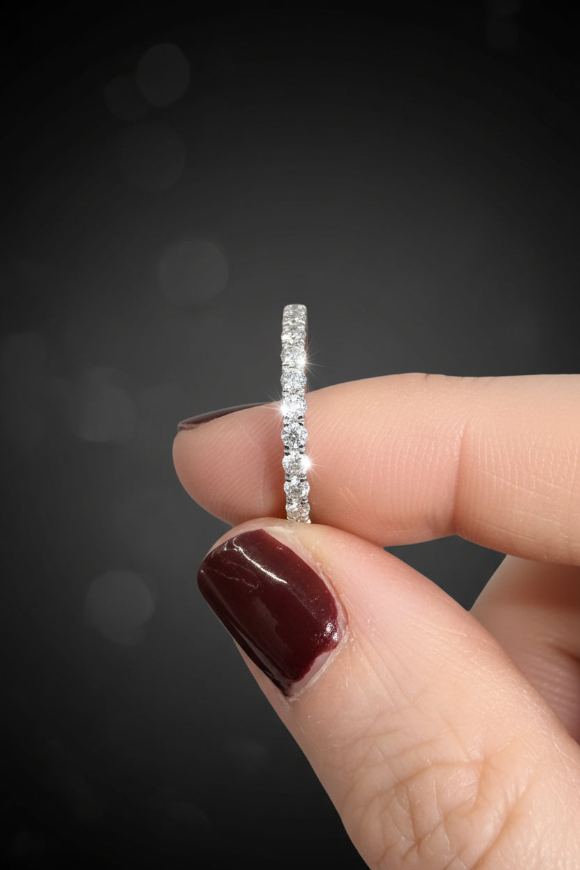 Product photograph of a brilliant diamond eternity band held between fingers with glossy burgundy nails against a dark studio background featuring sparkling light flares and soft bokeh circles