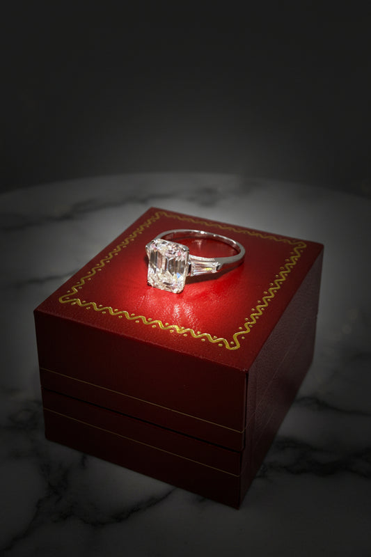 Photograph of a Cartier-style emerald-cut diamond engagement ring with tapered baguette side stones resting on a signature red jewelry box featuring gold scrollwork atop a white marble surface under a dramatic spotlight