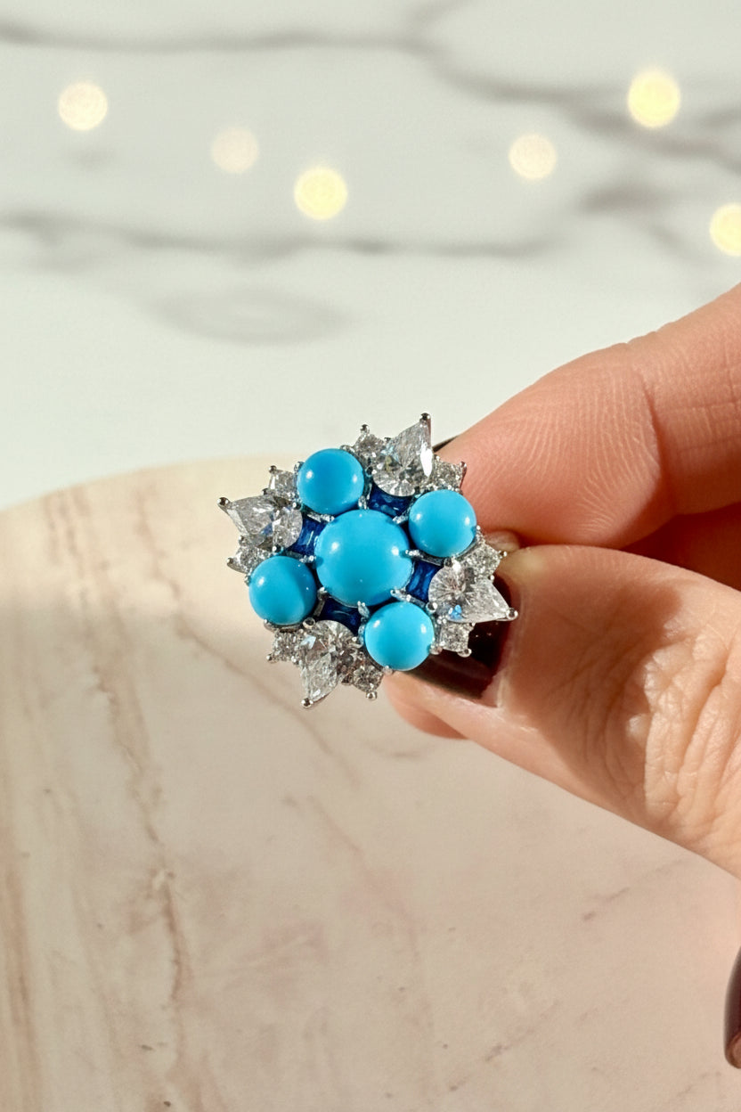 A photograph of a high-jewelry ring held by a hand featuring a central turquoise cabochon as a focal point complemented by brilliant pear-cut diamonds and vibrant blue sapphire accents against a blurred bokeh background