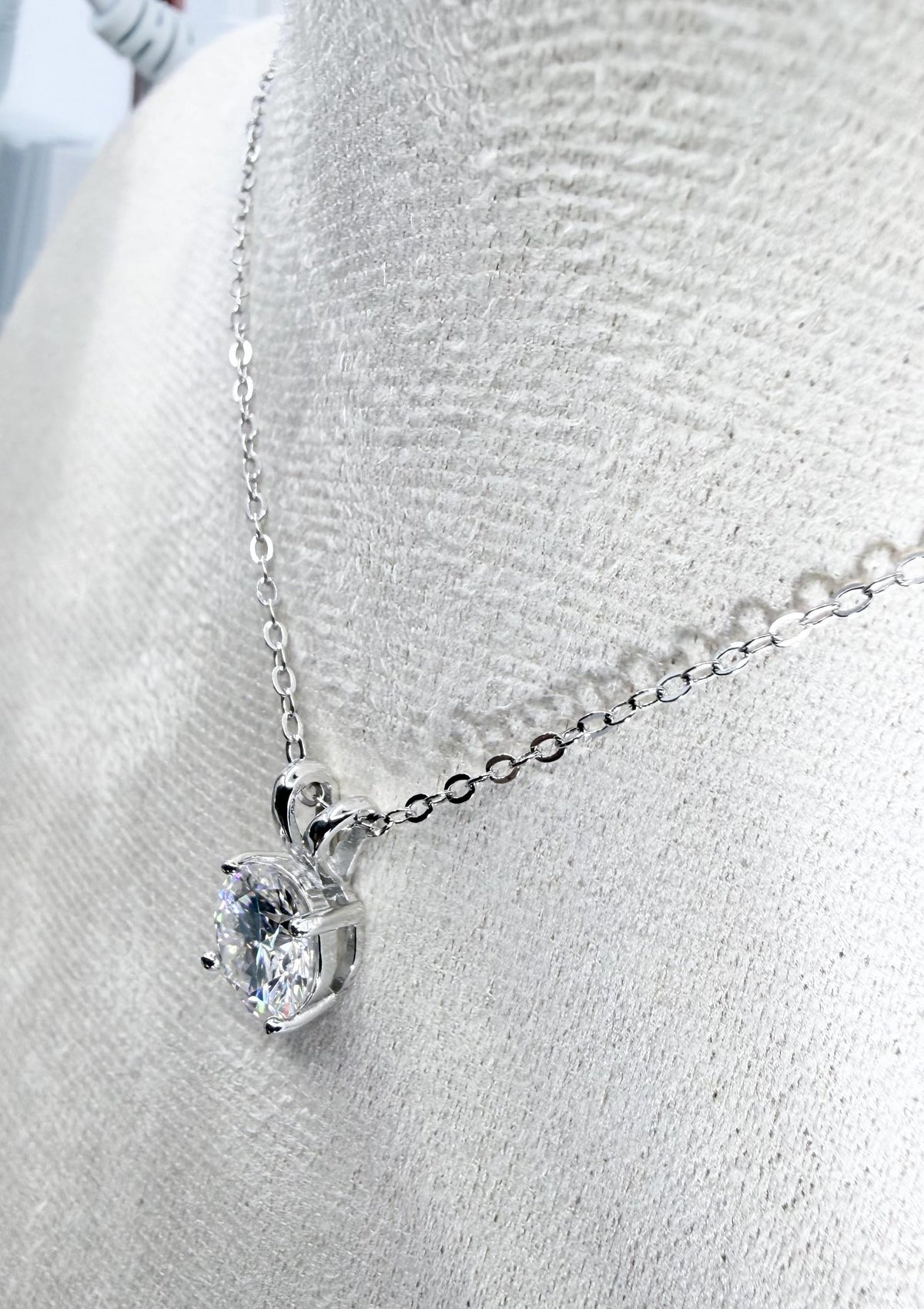 Professional jewelry photograph of a brilliant round-cut solitaire pendant necklace displayed on a light-colored textured bust featuring a polished four-prong setting, a shimmering silver cable chain, and intricate light-catching facets