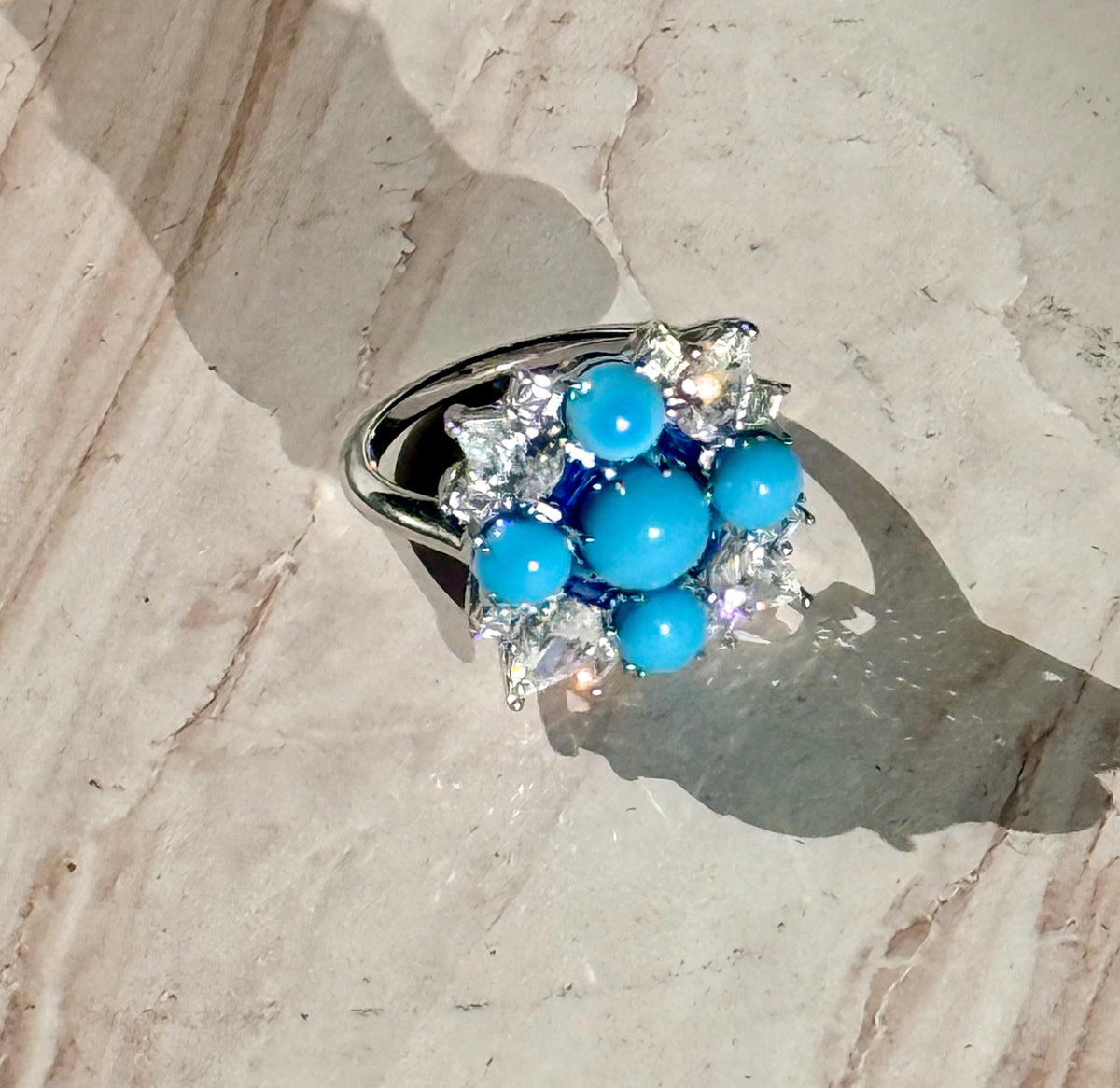 Fine jewelry silver ring featuring a central cluster of five vibrant sky-blue turquoise cabochons and shimmering white accent stones resting on a textured beige marble surface