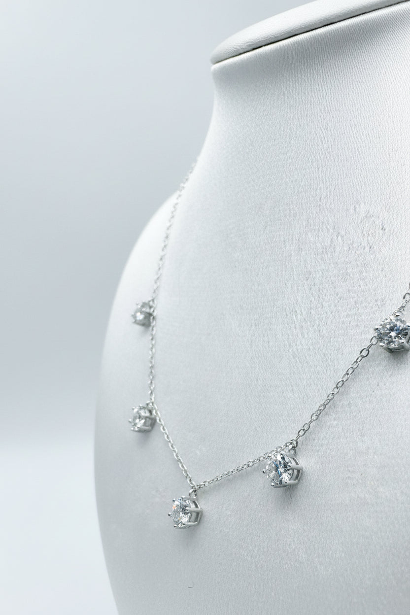 Photograph of a silver necklace featuring five dangling prong-set round brilliant stones displayed on a white jewelry bust with a clean high-key aesthetic