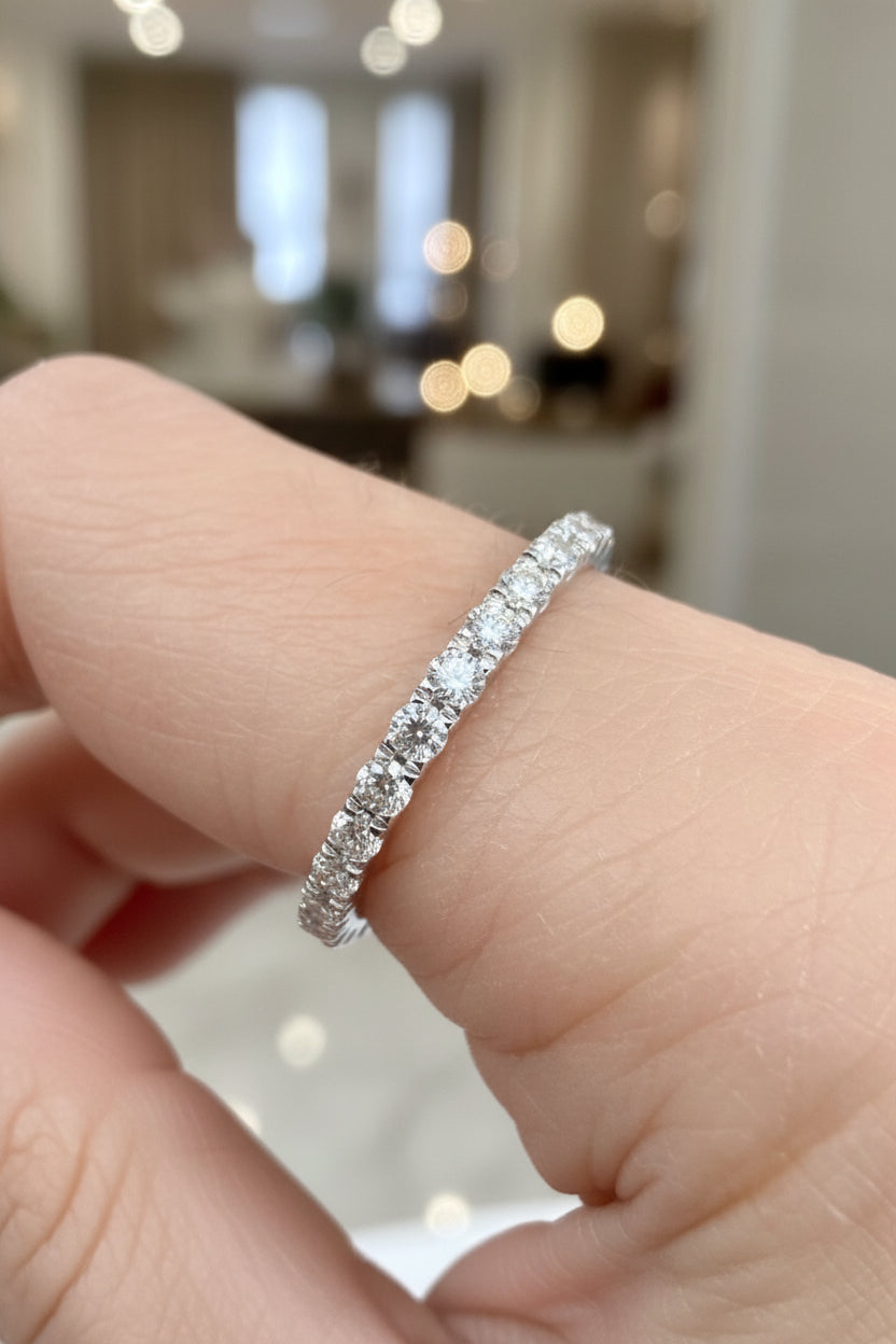 Digital photograph of a round-cut diamond eternity wedding band on a person's finger in a jewelry showroom featuring shimmering brilliant diamonds, a shared-prong white gold setting, and a warm blurred bokeh background