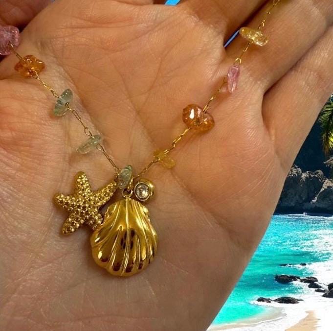 Gold-toned charm necklace held in a hand over a tropical beach background with turquoise water and rocky cliffs, featuring a textured gold starfish, a polished seashell charm, and multicolored raw gemstone beads