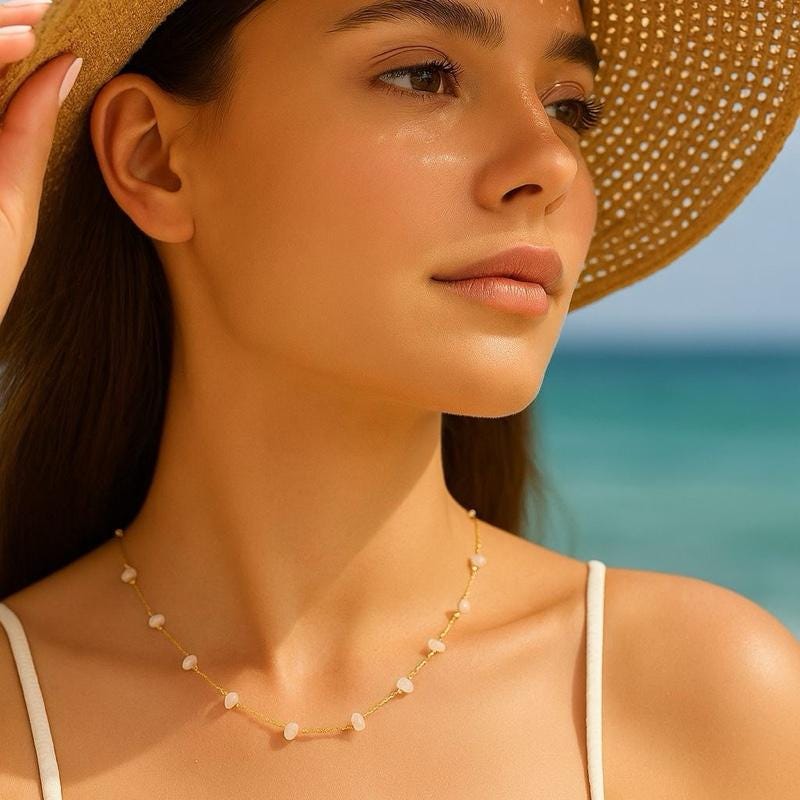 Digital lifestyle photograph of a young woman at the beach wearing a woven straw sun hat and a delicate gold-toned necklace with small white beads against a blurred turquoise ocean horizon