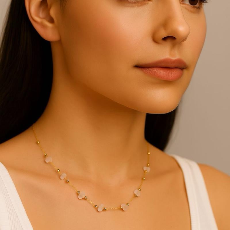 Close-up photograph of a woman in a studio setting wearing a delicate gold chain necklace featuring pale pink heart-shaped stones and small gold beads while wearing a white ribbed tank top