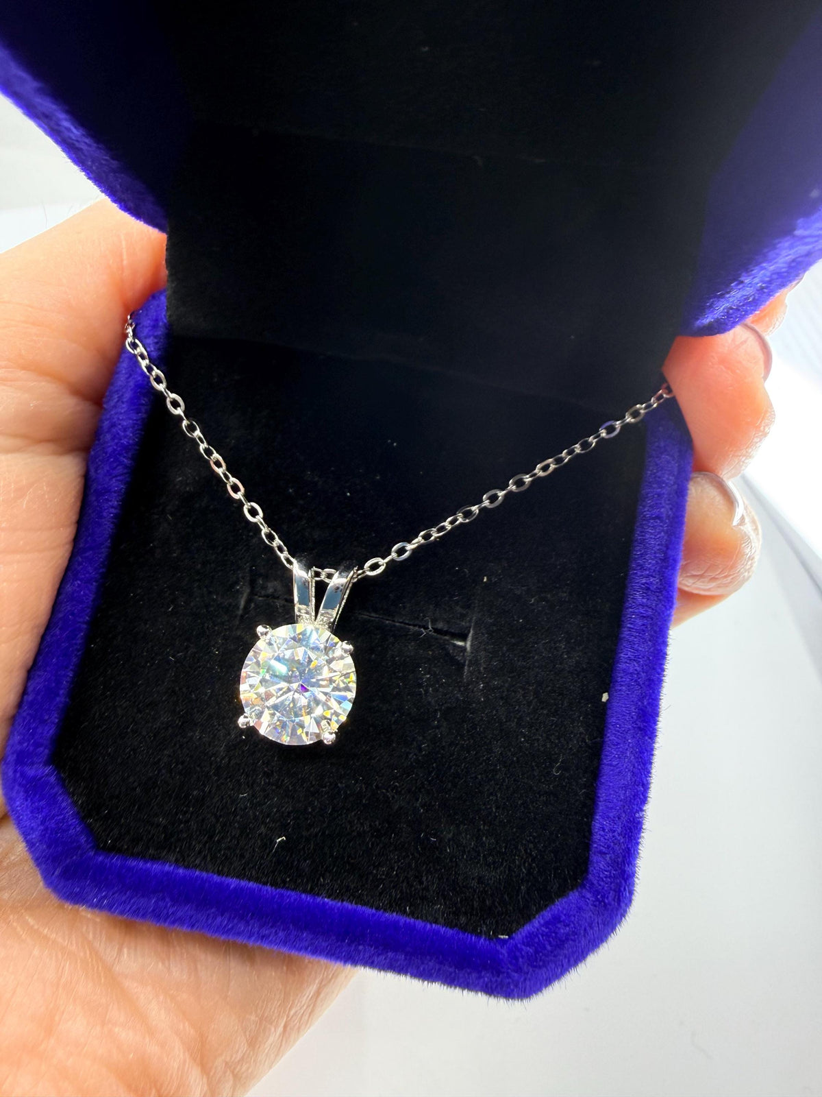 Photograph of a round brilliant cut solitaire pendant necklace nestled in a royal blue velvet jewelry box held by a hand, highlighting the prismatic rainbow fire of the stone and its delicate silver setting and chain