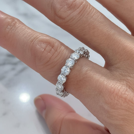 Close-up photograph of a round brilliant-cut diamond eternity band worn on a finger over a white marble surface featuring shimmering white gemstones, a silver-toned shared-prong setting, and a bright reflected light flare