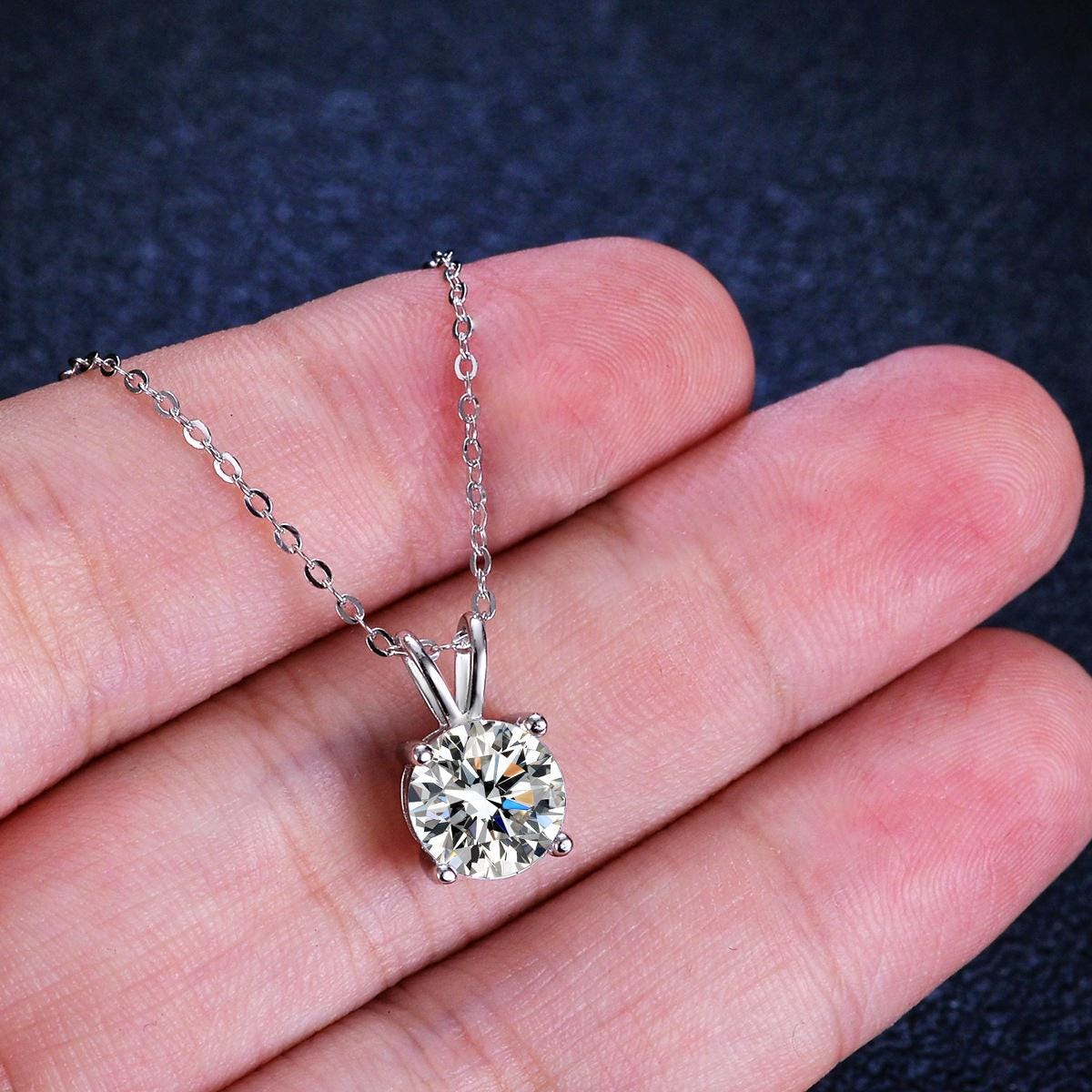 Photograph of a round brilliant-cut diamond solitaire necklace held in a hand featuring a sparkling multi-faceted clear gemstone in a four-prong silver setting and a delicate cable chain