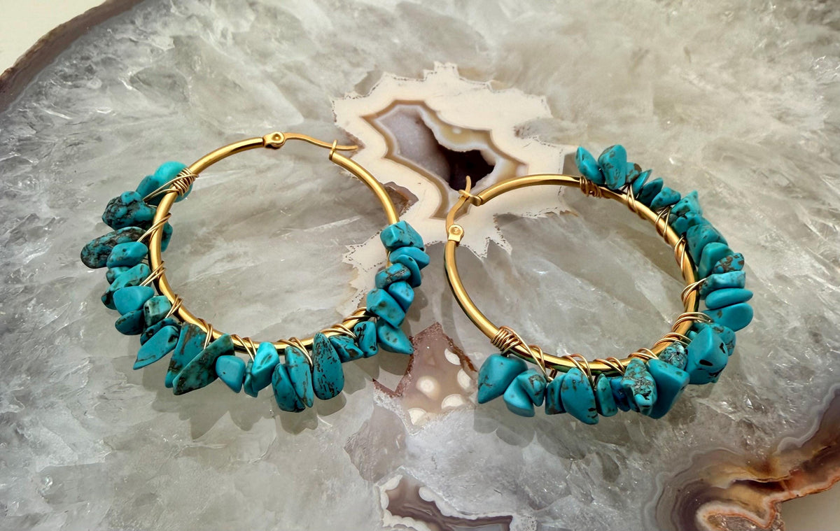Handmade wire-wrapped jewelry featuring a pair of gold hoop earrings with raw vibrant turquoise stone chips and dark matrix patterns displayed on a translucent white crystalline geode slice