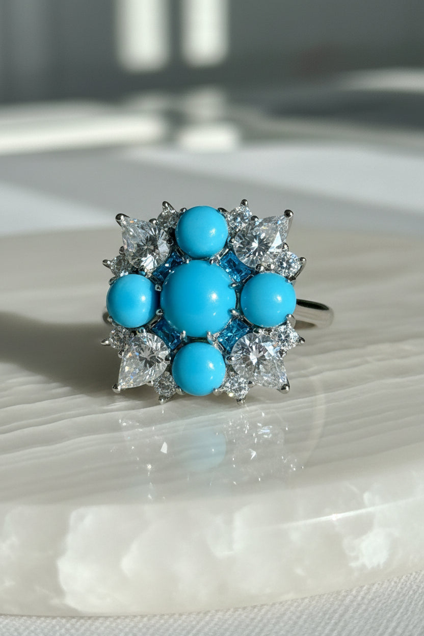 Fine jewelry turquoise and diamond cocktail ring resting on a white rippled stone surface featuring five vibrant azure cabochons and brilliant pear-cut gems in a symmetrical sunburst arrangement
