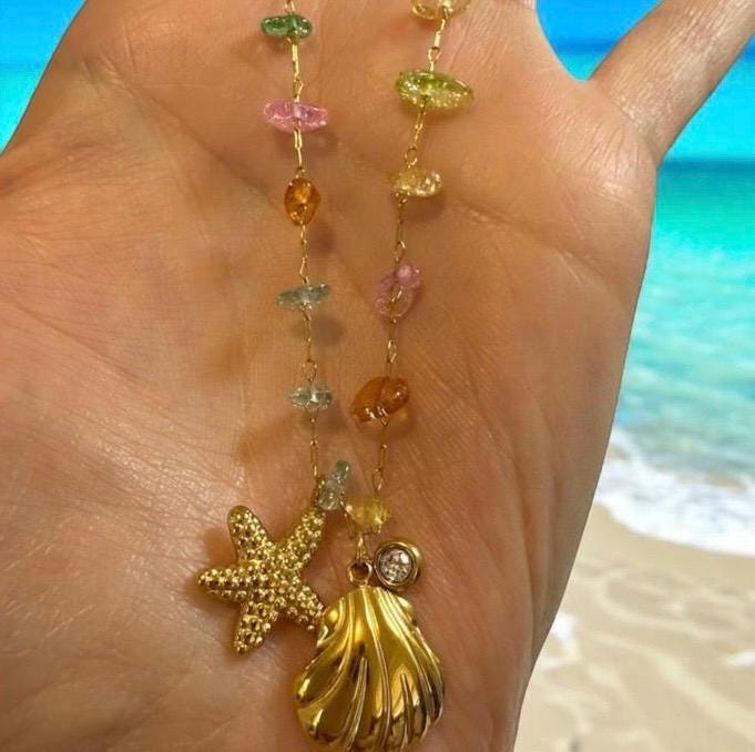 Close-up photograph of a gold necklace held in an open hand at a tropical beach featuring a textured starfish charm, a ribbed seashell pendant, and a delicate chain adorned with translucent multi-colored gemstone beads in shades of pink, green, and amber