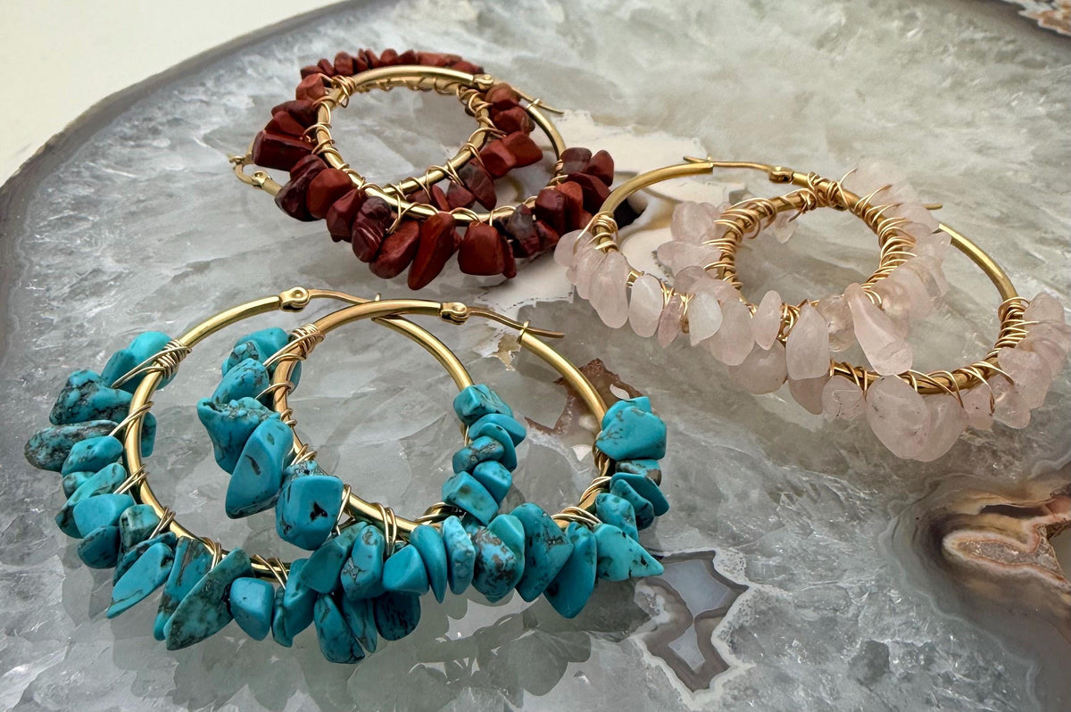 Handmade wire-wrapped jewelry featuring three pairs of gold hoop earrings embellished with vibrant turquoise, translucent rose quartz, and deep red jasper chips resting on a polished crystalline agate slab