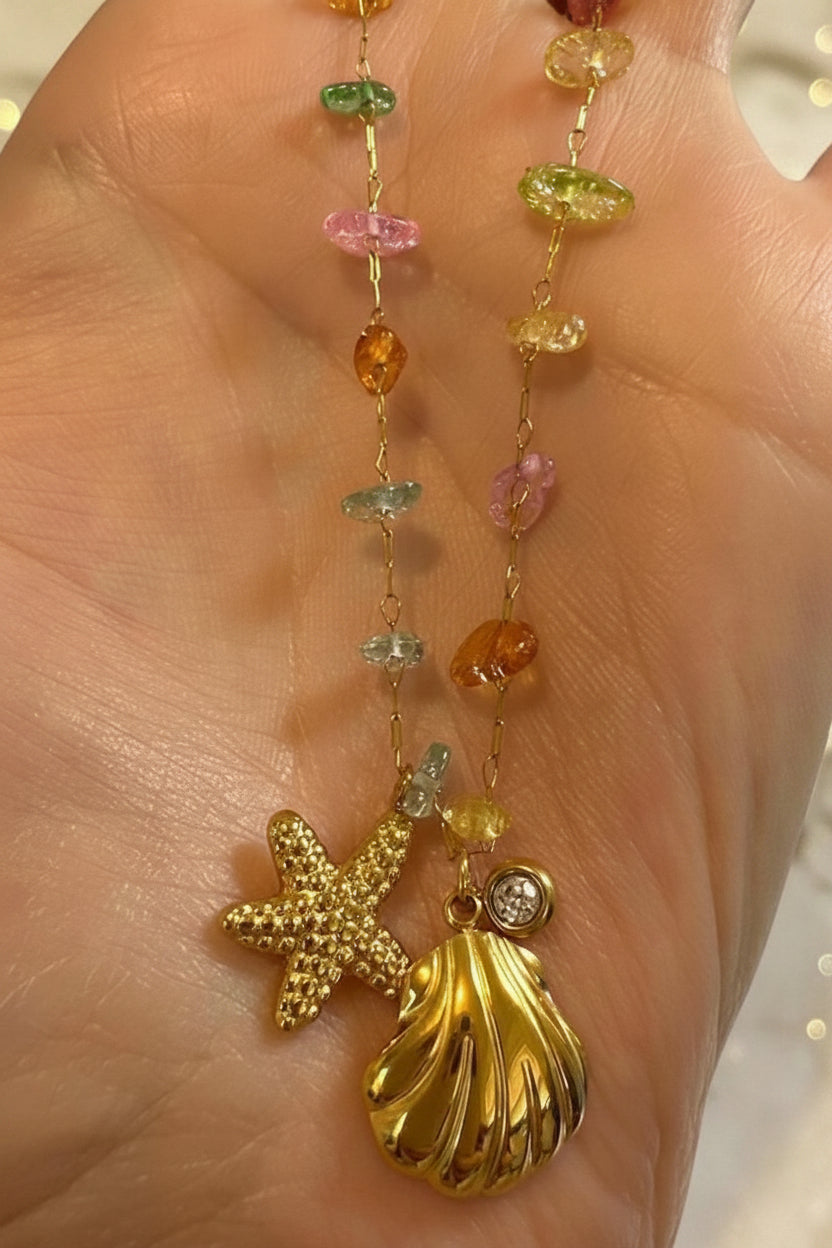 Gold charm necklace held in a person's palm featuring a textured starfish and polished seashell with a small diamond accent along a chain of colorful gemstone beads in pink, green, and orange hues