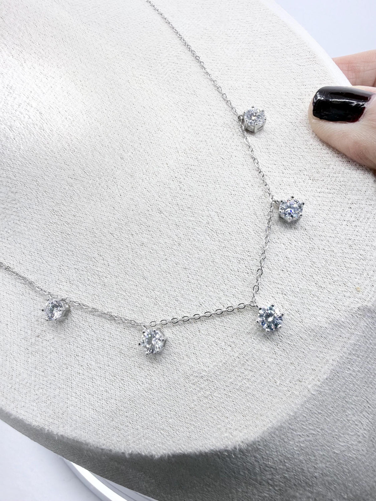 Silver jewelry piece featuring a dainty necklace with five brilliant-cut clear stones in prong settings displayed on a textured white bust held by a finger with glossy black nail polish