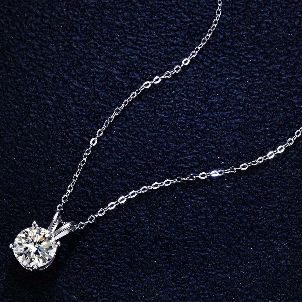 Photograph of a round brilliant-cut diamond pendant necklace resting on a textured navy blue fabric featuring a four-prong silver setting and a delicate shimmering cable chain
