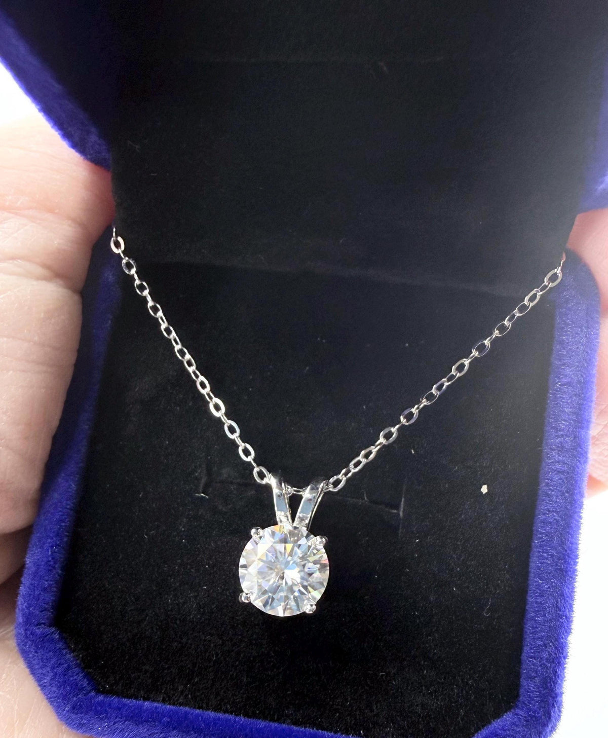 Photograph of a brilliant round-cut diamond solitaire pendant on a silver chain nestled in a royal blue velvet jewelry box held by a hand to showcase the sparkling facets and delicate four-prong setting