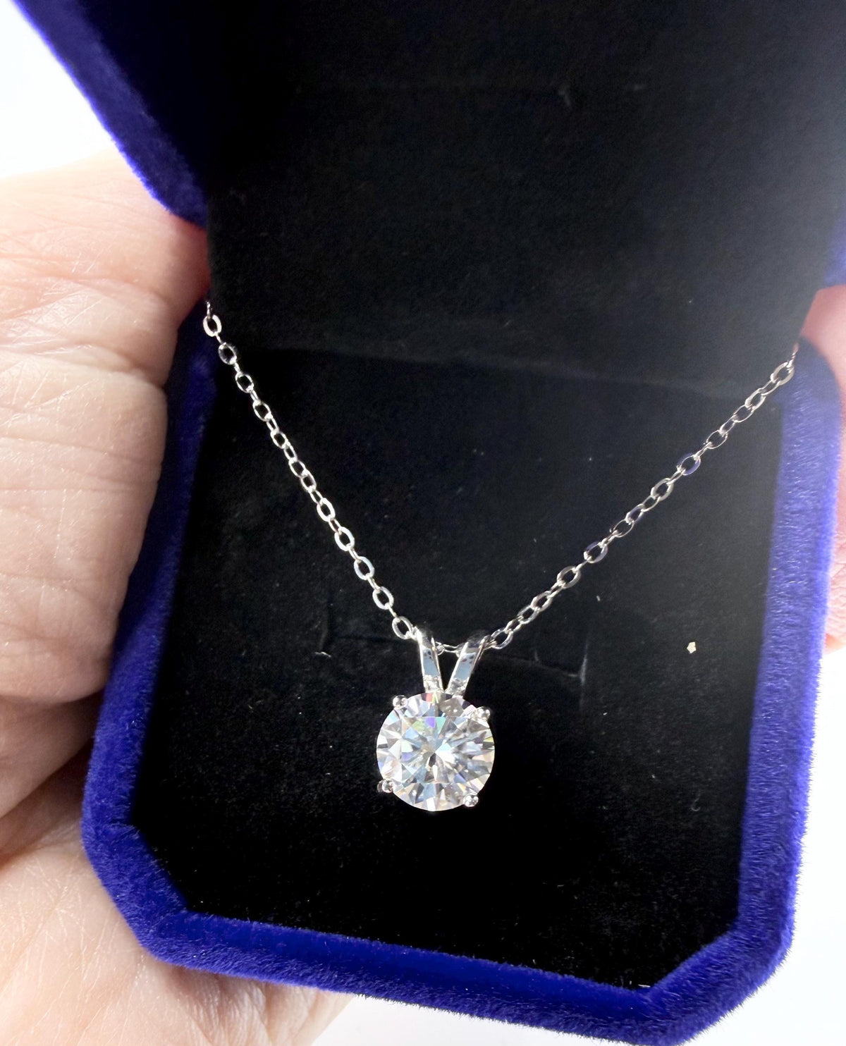 Photograph of a brilliant round-cut diamond solitaire necklace on a silver chain displayed in a royal blue velvet box with a plush black interior and sparkling prismatic light reflections within the faceted stone