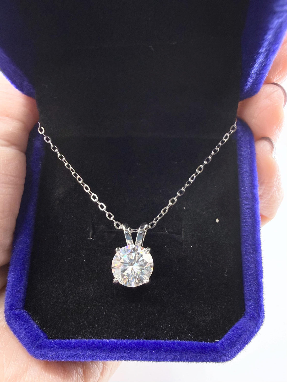 Photograph of a brilliant round-cut solitaire pendant necklace nestled in a plush royal blue velvet jewelry box with a black lining featuring a delicate silver chain and prismatic light reflections within the gemstone facets
