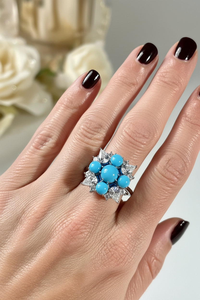 Photograph of a vibrant turquoise and diamond cluster ring on a hand with dark burgundy nail polish, featuring a central round cabochon surrounded by smaller turquoise stones and shimmering pear-cut diamonds set against a soft white floral backdrop