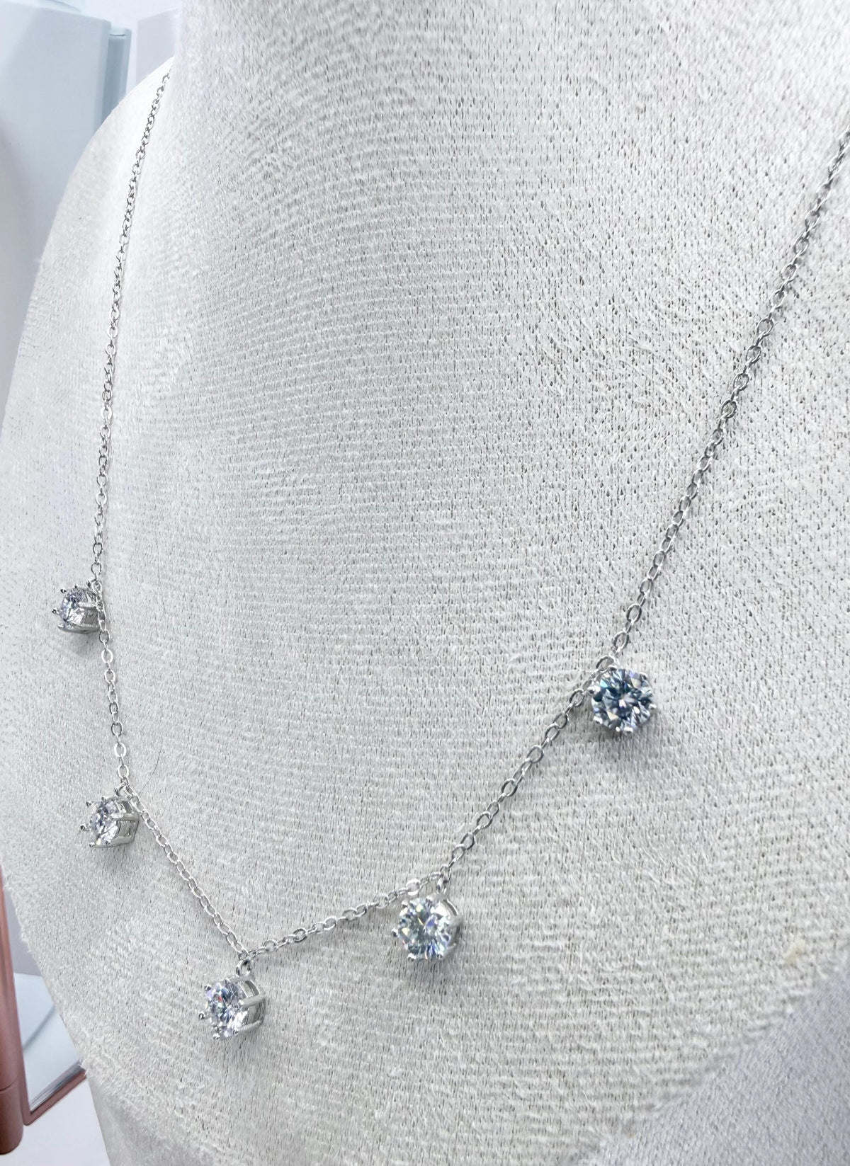 Silver-tone jewelry necklace featuring five round-cut brilliant gemstones in delicate prong settings displayed on a light gray textured fabric mannequin bust with a thin cable chain and shimmering facets