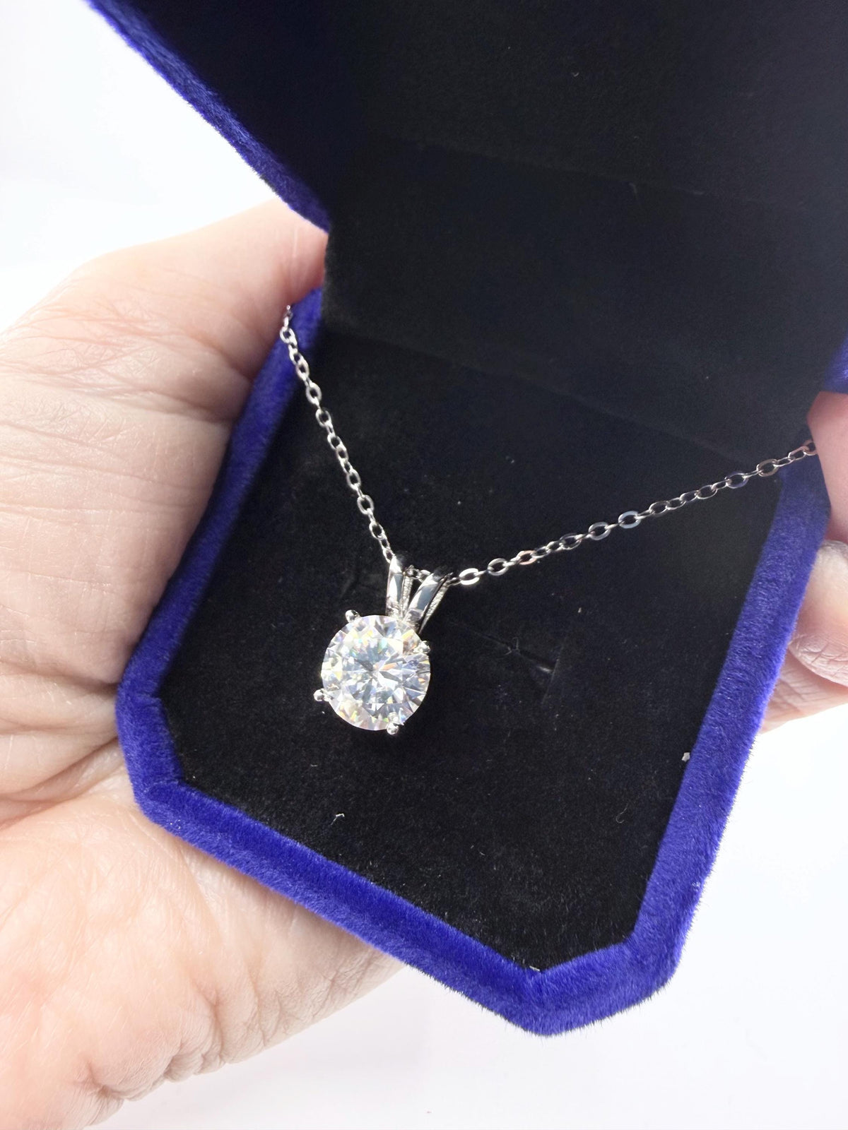 Photograph of a silver solitaire pendant necklace featuring a large round brilliant-cut clear gemstone showcased in an open royal blue velvet jewelry box with a black interior lining held by a hand