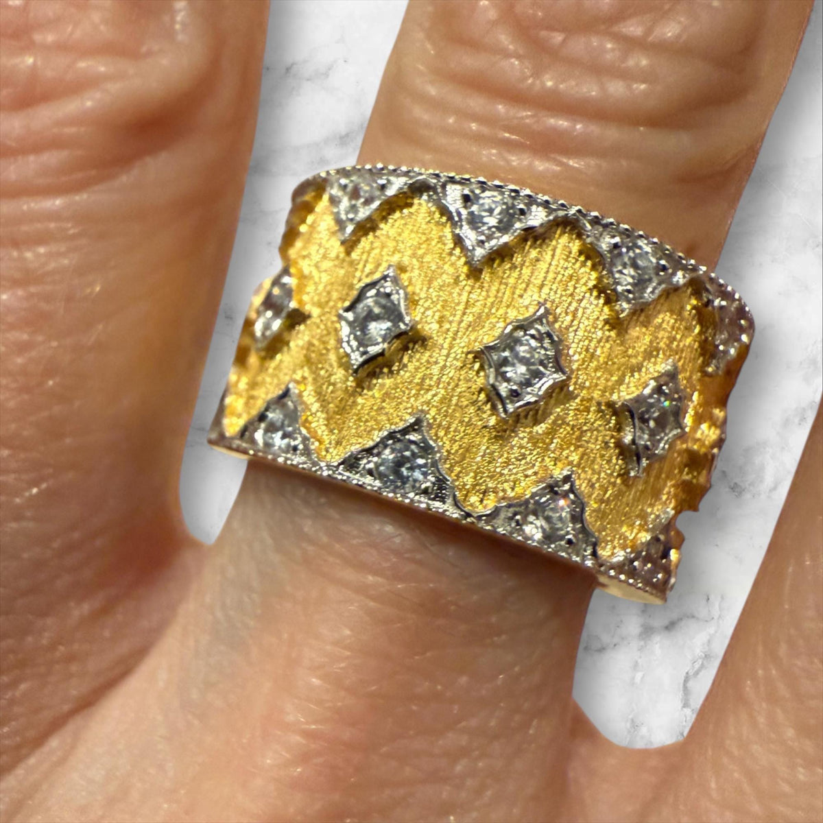 Hand-engraved 18k yellow and white gold diamond wide band ring by Buccellati worn on a finger featuring a brushed rigato texture, scalloped diamond-set borders, and central rhombus-shaped diamond accents against a marble background