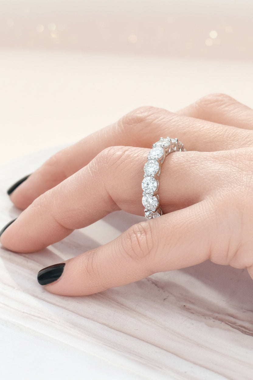 Digital photograph of a round brilliant-cut diamond eternity ring worn on a hand with glossy black nails resting on a light-veined marble surface with a soft golden bokeh background