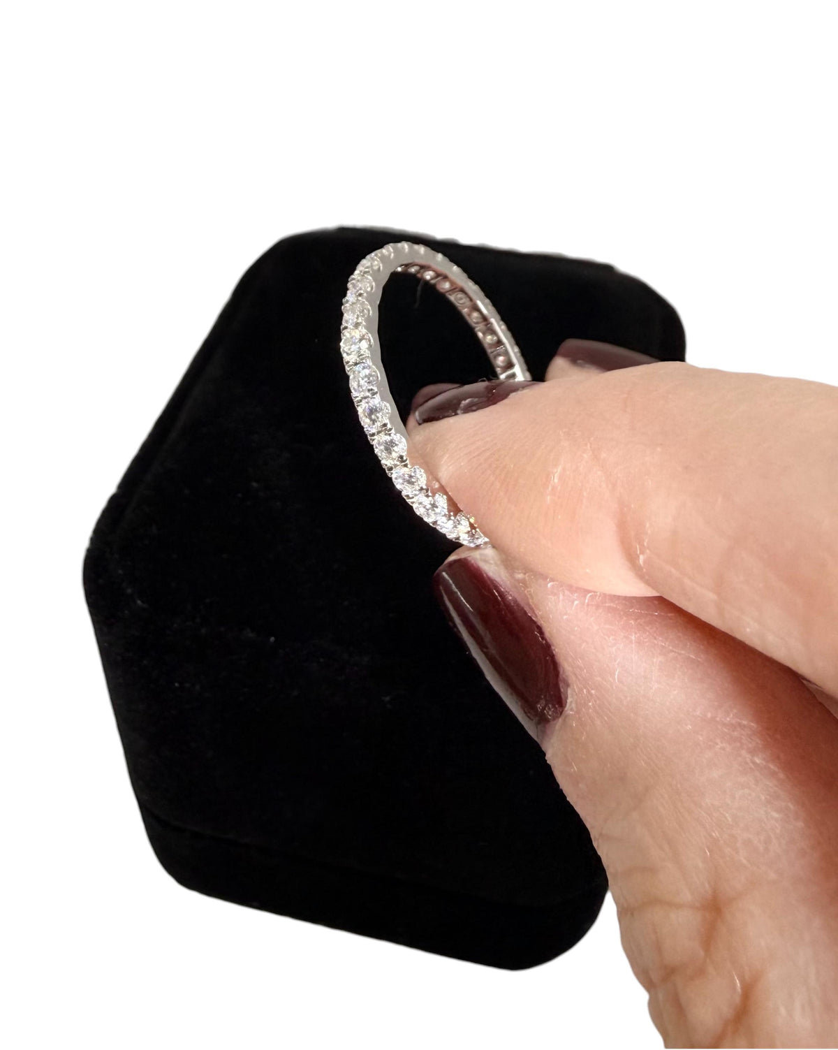 Photograph of a brilliant-cut diamond eternity ring held by a hand with maroon nail polish over a black velvet jewelry box against a clean white background