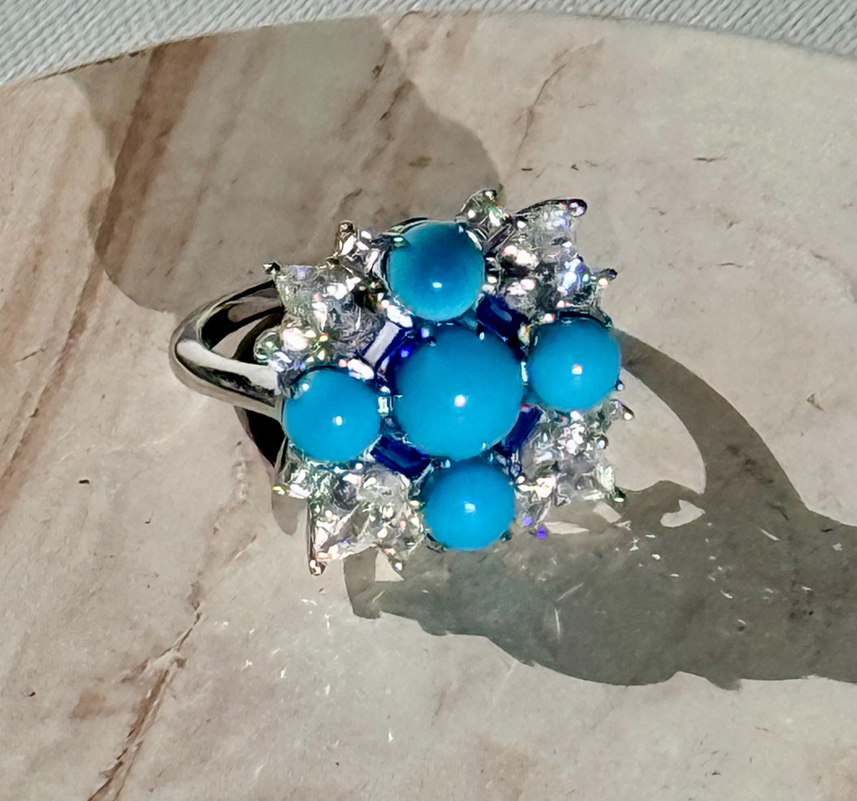 Handcrafted gemstone ring featuring five vibrant turquoise cabochons and shimmering diamond accents in a starburst cluster resting on a neutral stone surface