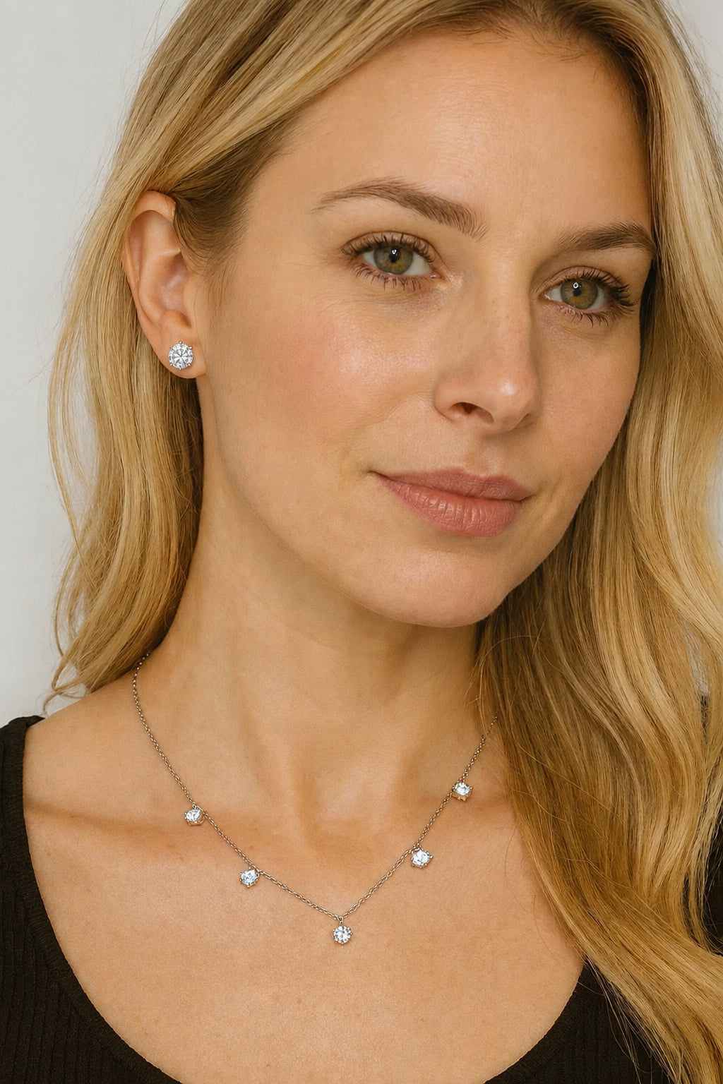 Photograph of a blonde woman in a studio setting wearing a delicate silver necklace with five round-cut diamond pendants, highlighting her green eyes and a black V-neck shirt