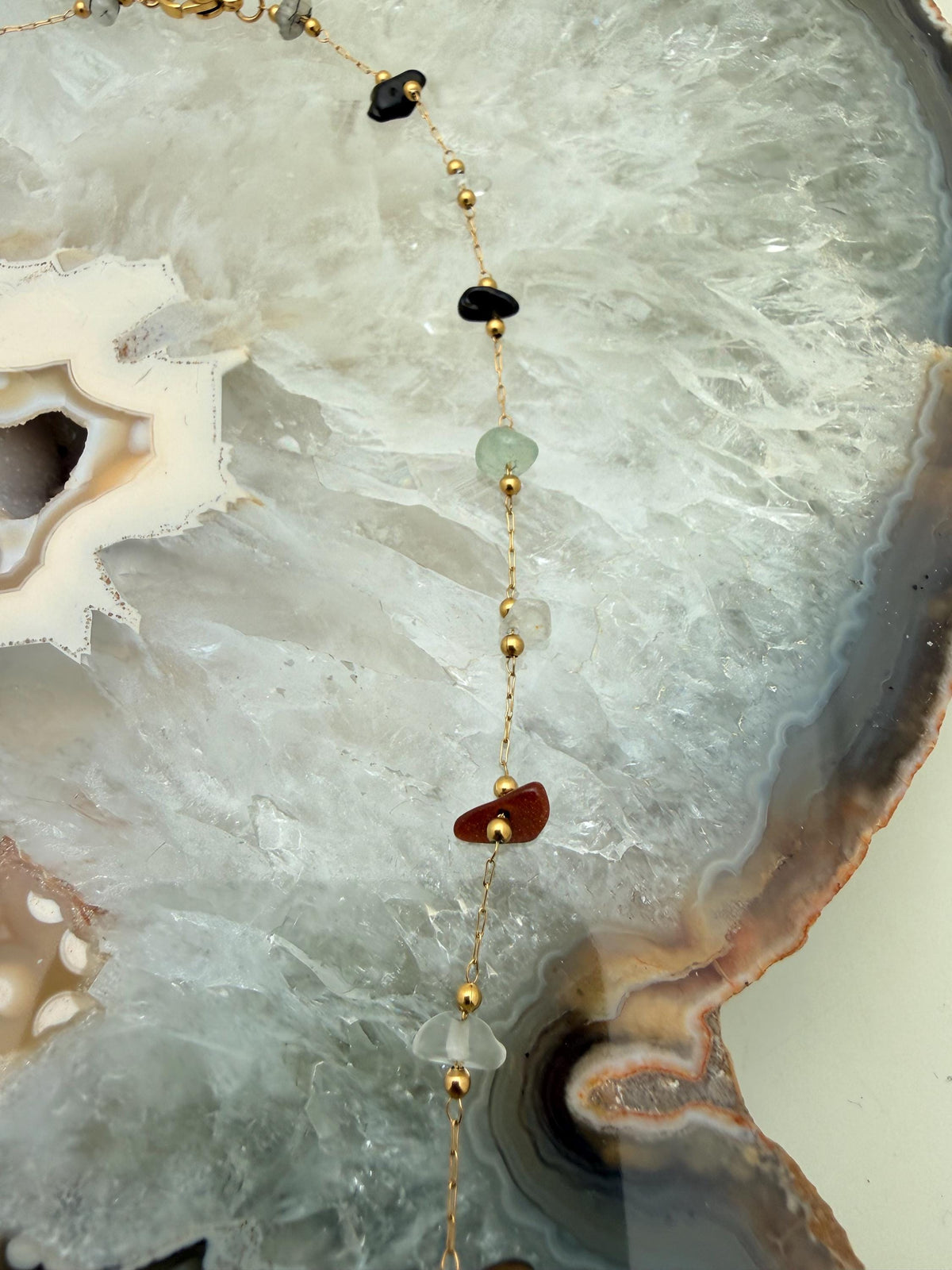 Photograph of a handmade gold necklace with gemstone chips draped over a white crystalline agate geode slice featuring small gold beads, a sparkling reddish-brown goldstone, and a pale green stone chip